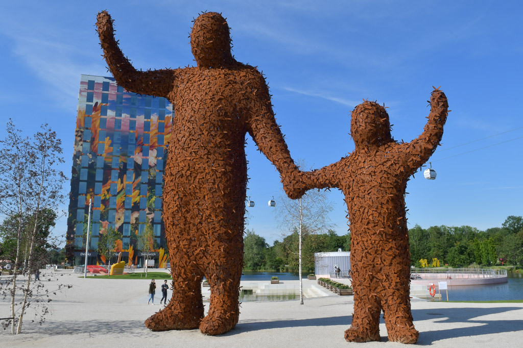 Skulptur aus Zweigen: Riese mit Kind vor einem bunten Hochhaus