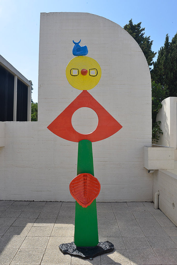 Skulptur in Rot, Grün und Gelb vor weißer Mauer