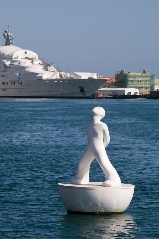 Weiße Skulptur einer Figur auf einem Sockel im Hafenwasser